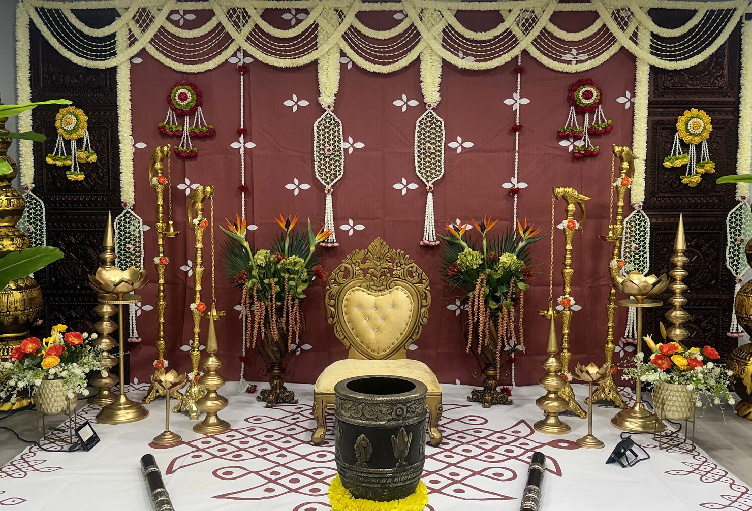Traditional ceremony stage with a maroon backdrop, jasmine garlands, carved gold pillars, brass lamps and a marigold-wrapped mortar and pestles
